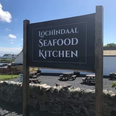 The slate Lochindaal Seafood Kitchen sign, with picnic benches and a drystone wall behind